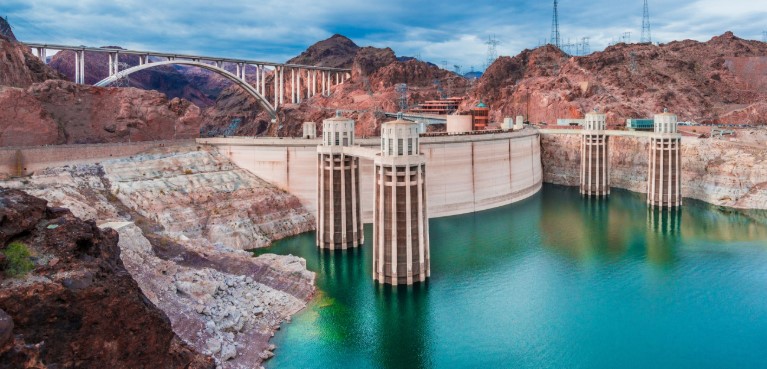 Photo is of Hoover Dam. The water is beautiful and shows a reflection of the dam. The bridge is also shown in the top of the image.